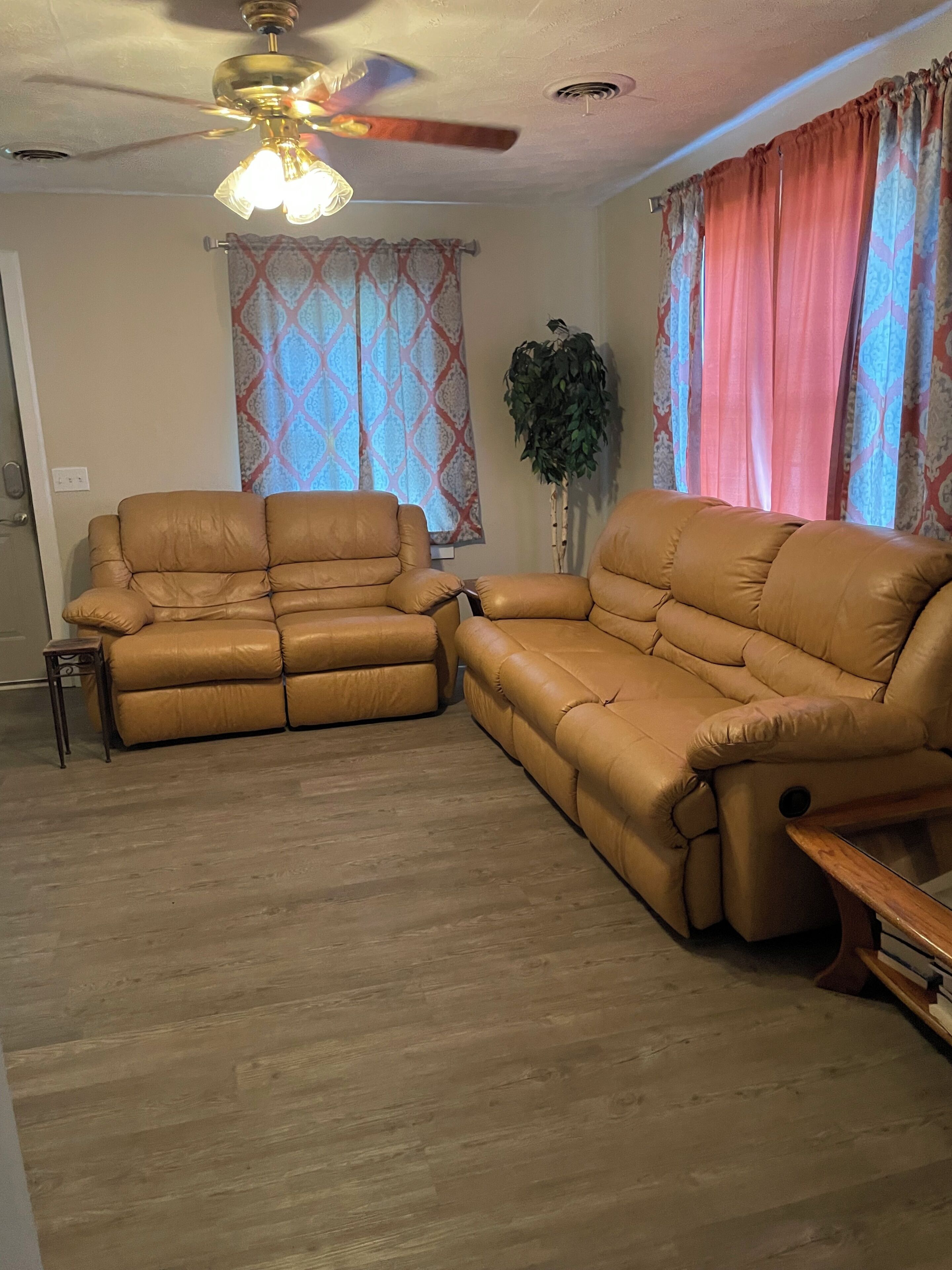 Una sala de estar con un sofá de cuero marrón, un ventilador de techo, cortinas estampadas y una planta en maceta.
