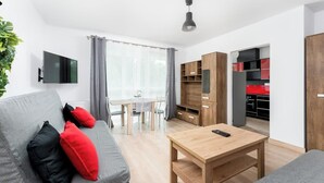 Een moderne woonkamer met een bank, een salontafel en een televisie. Er is een eethoek met een tafel en stoelen. De kamer heeft houten kasten en een keuken in de achtergrond.