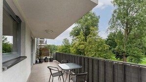 Een balkon met een tafel en stoelen, een plant in een pot, en uitzicht op bomen en een gebouw op de achtergrond.