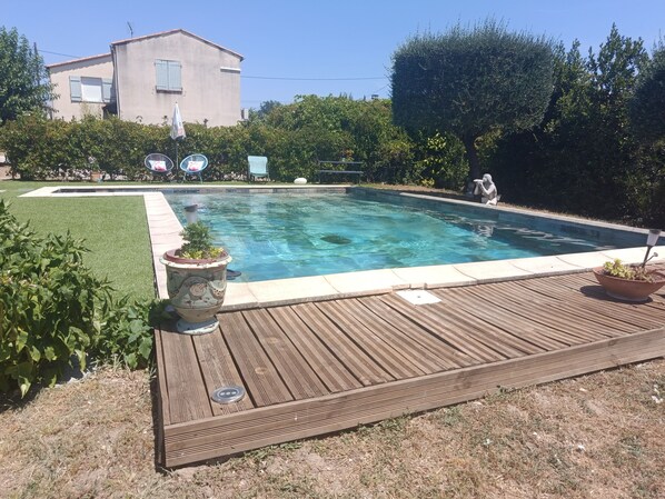 En swimmingpool med en træterrasse, omgivet af grøn vegetation og en bygning i baggrunden.