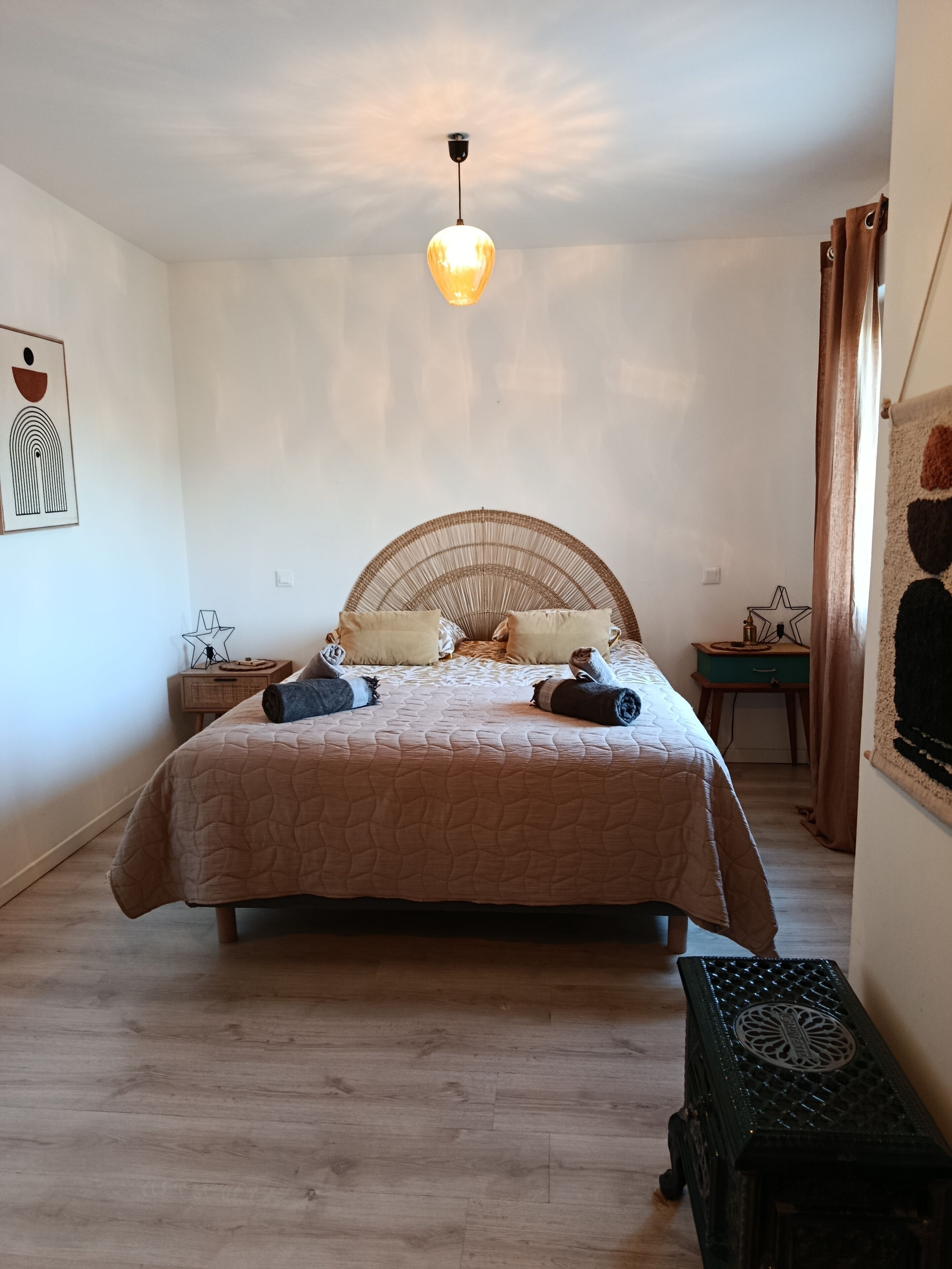Un dormitorio con cama, lámpara colgante, mesita de noche, un taburete pequeño y un adorno de pared.
