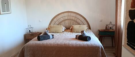 A bedroom with a bed, a hanging light fixture, a bedside table, a small stool, and a wall decoration.