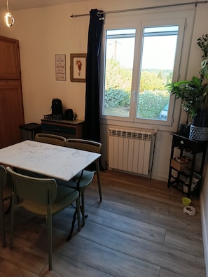 A room with a dining table, chairs, a radiator, a window with a view of greenery, and a plant on a shelf. - Time Regained (Saint-Martin-de-Valgalgues)