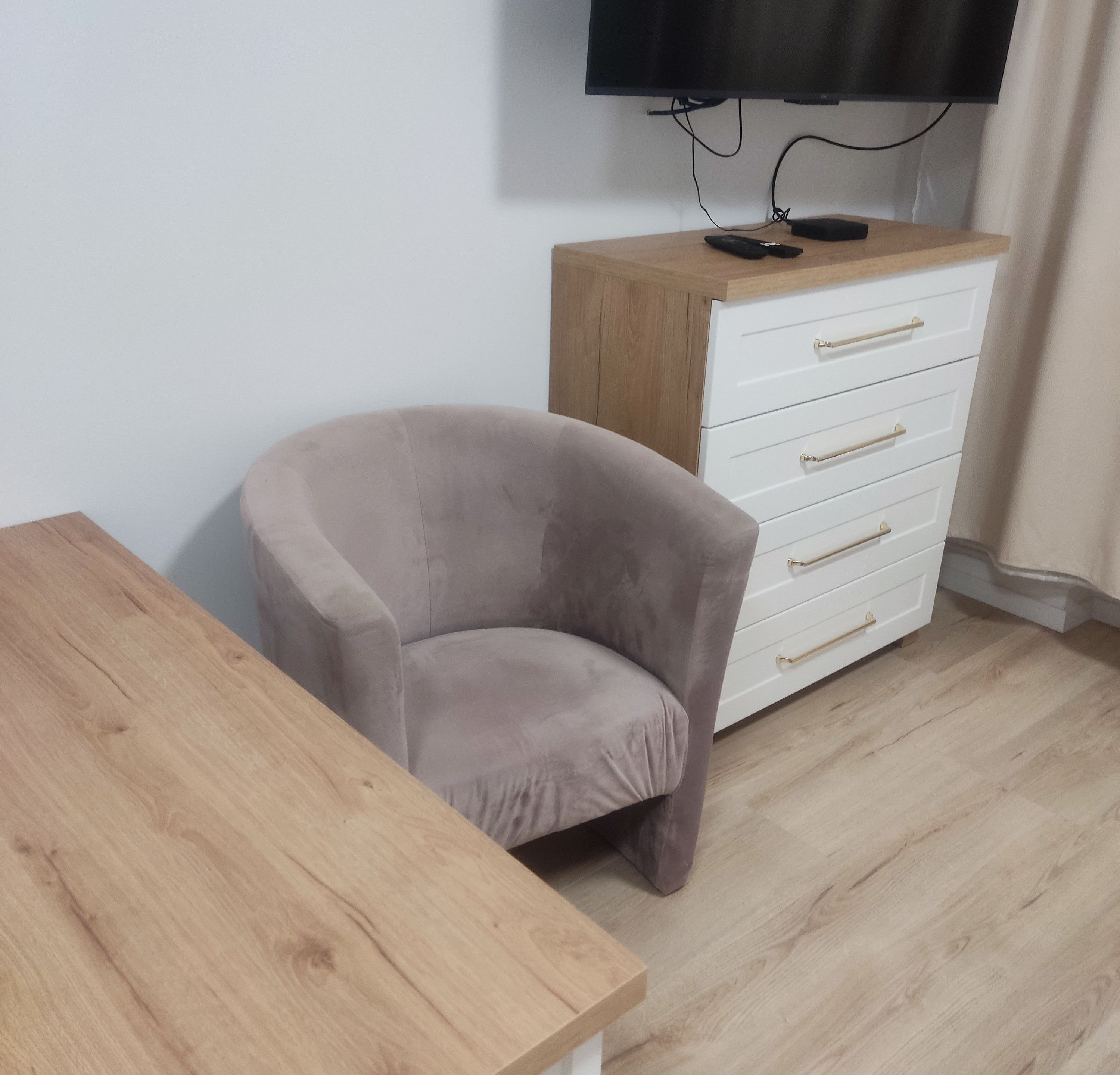 A room with a wooden table, a grey armchair, a white dresser with drawers, and a television mounted on the wall.