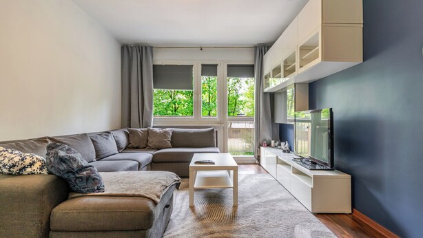 Un salon moderne avec un canapé d’angle, une petite table blanche, un téléviseur et une vue sur la verdure à l’extérieur.