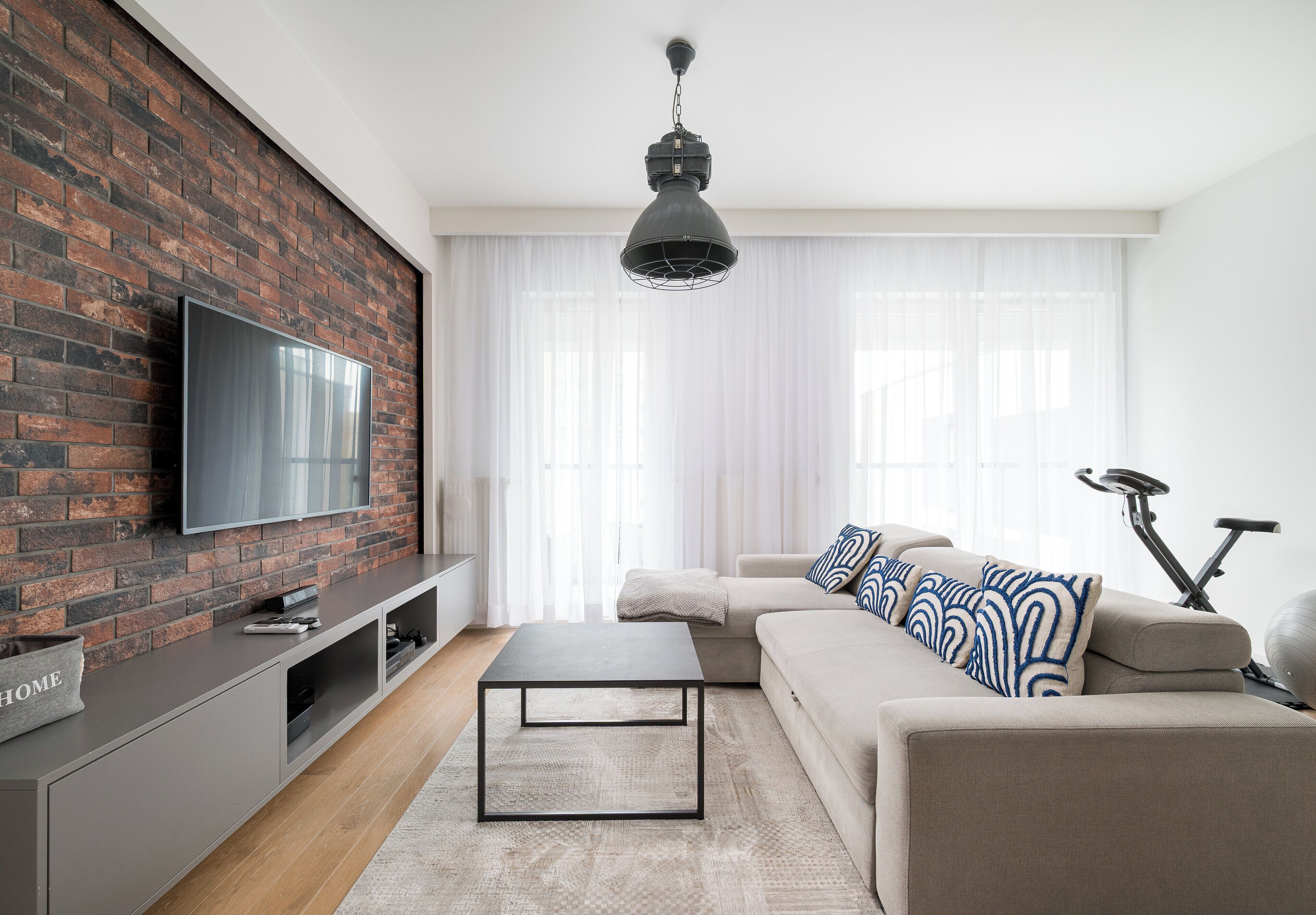 A modern living room with a brick wall, flat-screen TV, sofa, coffee table, and exercise bike.