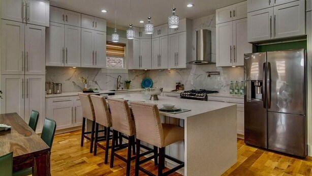 Una cocina moderna con armarios blancos, una isla central con taburetes de bar, un refrigerador de acero inoxidable e iluminación colgante.