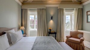 Habitación de hotel con una cama grande, dos ventanas con cortinas, una mesita de noche con lámpara y una silla.