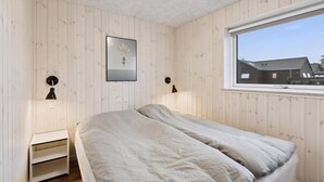 Ein Zimmer mit Holzwänden, einem Bett mit weißer Bettdecke, zwei Nachttischlampen, einem kleinen weißen Nachttisch und einem Fenster mit Blick auf Häuser.