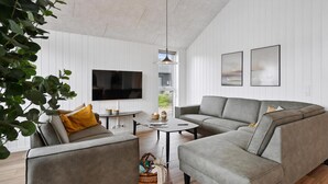 Ein modernes Wohnzimmer mit grauem Sofa, einem kleinen Couchtisch, einem an der Wand befestigten Fernseher und einem Fenster mit Blick ins Freie.