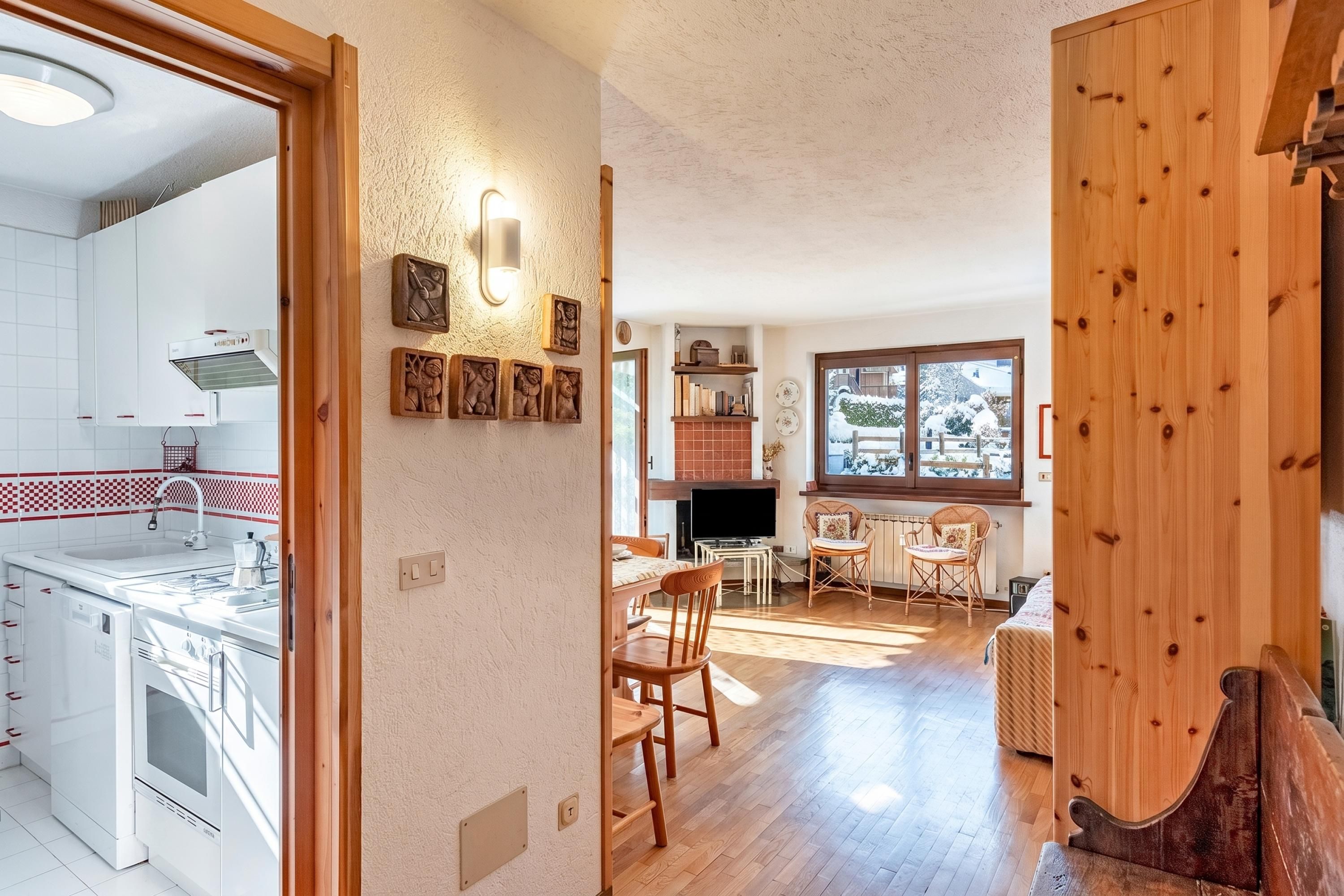 Una cocina moderna con armarios blancos, horno empotrado y un lavamanos. Hay un comedor con mesa y sillas, y una ventana con vistas al exterior.