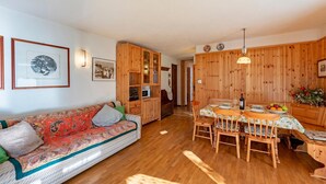 Ein gemütliches Zimmer mit einer Couch, Holzausbau, einem Esstisch mit Stühlen und einer Küchenzeile.