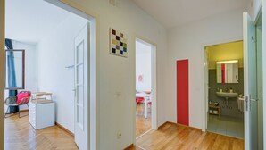 Una habitación con suelo de madera, una pared blanca con una obra de arte geométrica, una pared roja y una puerta de cristal que da al baño.