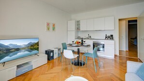 Un salon moderne doté d’un téléviseur à écran plat, d’un coin repas avec une table dressée pour deux personnes et d’une cuisine avec des meubles blancs.