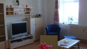 Ein Wohnzimmer mit Fernseher, blauem Sessel, Tisch mit Blumenvase und Regalen mit verschiedenen Gegenständen.