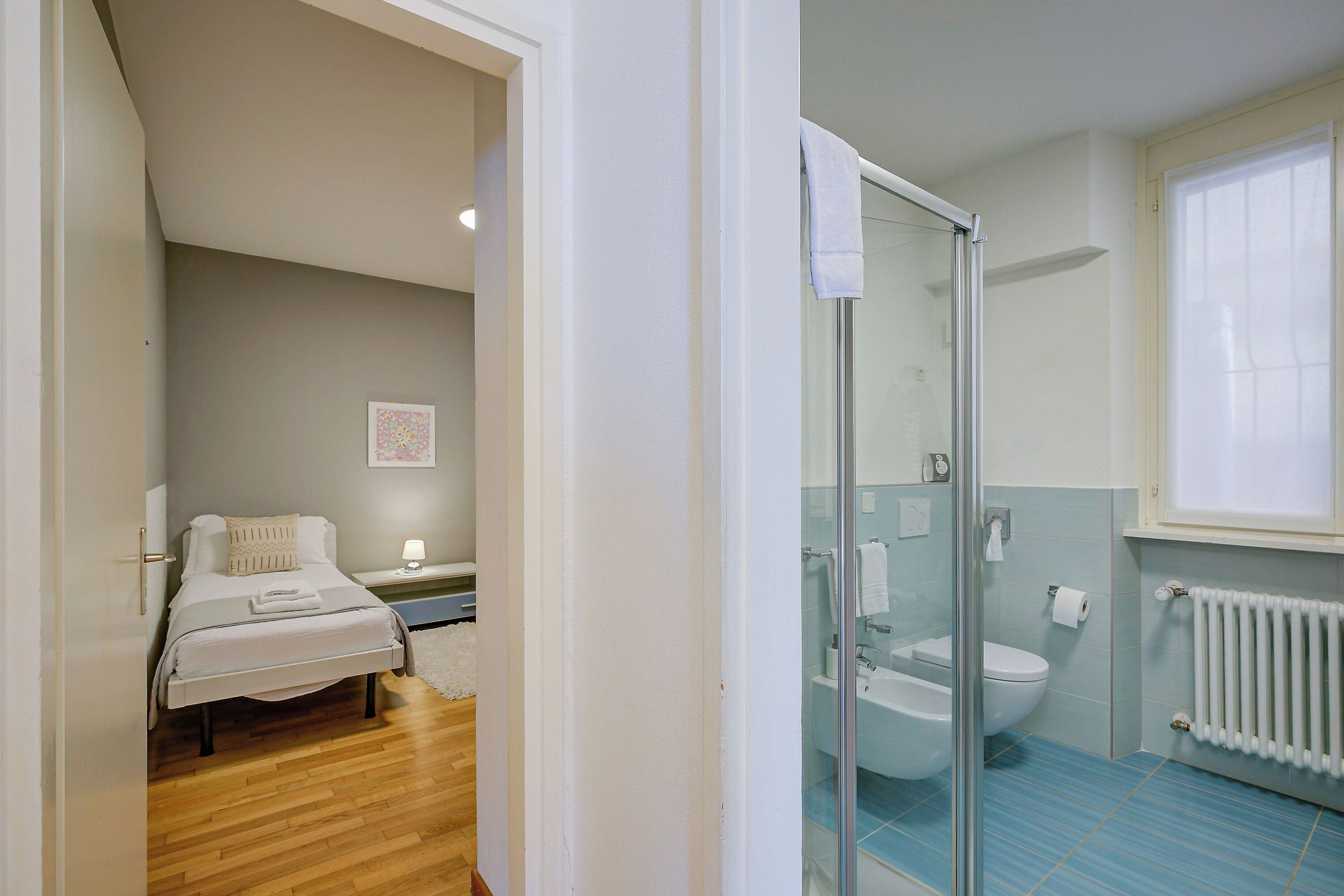 A bedroom with a bed, a bedside table, and a nightstand. There is a bathroom with a glass shower enclosure, a toilet, and a sink.