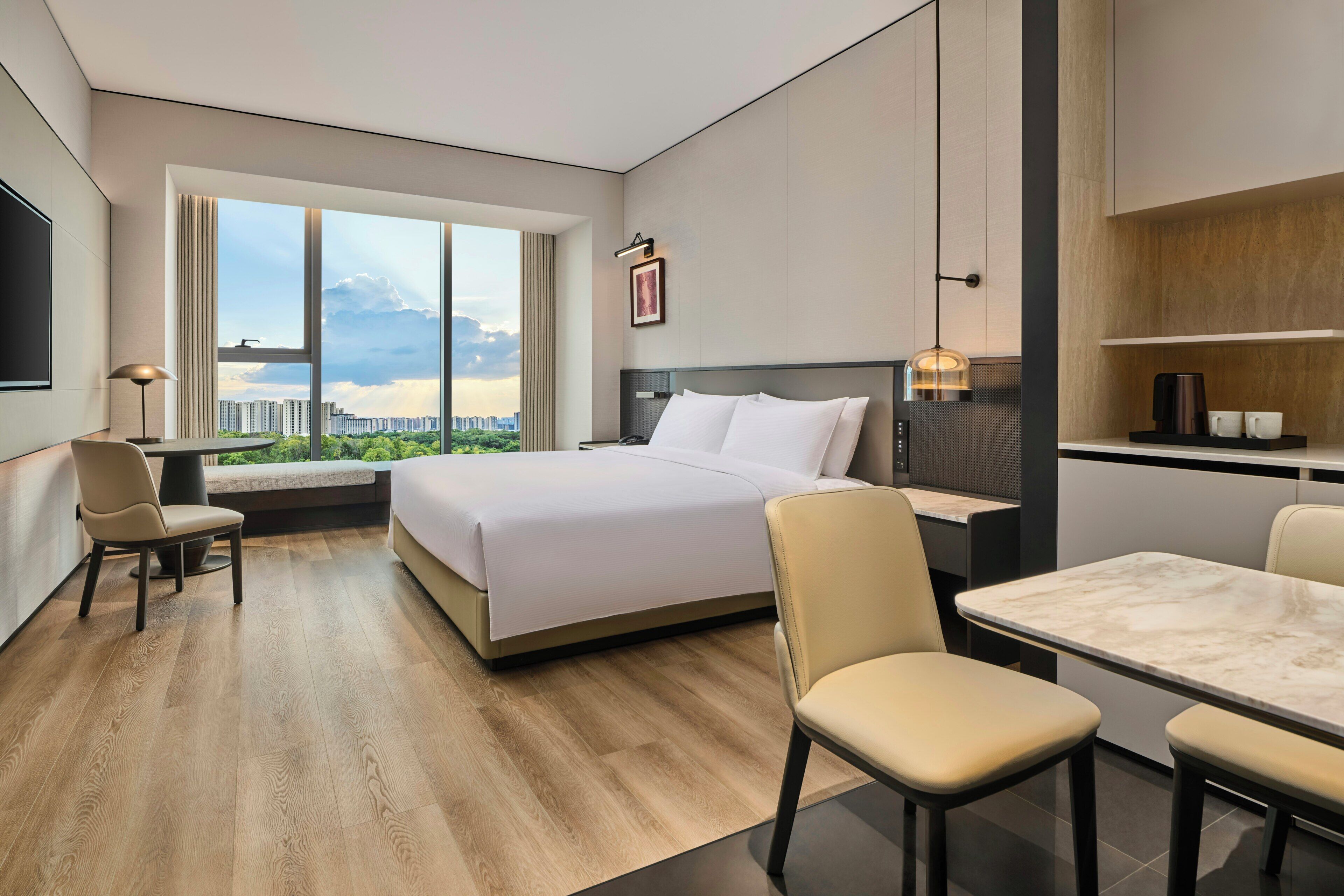 A hotel room with a bed, a desk, a chair, a dining table, and a large window offering a city view.