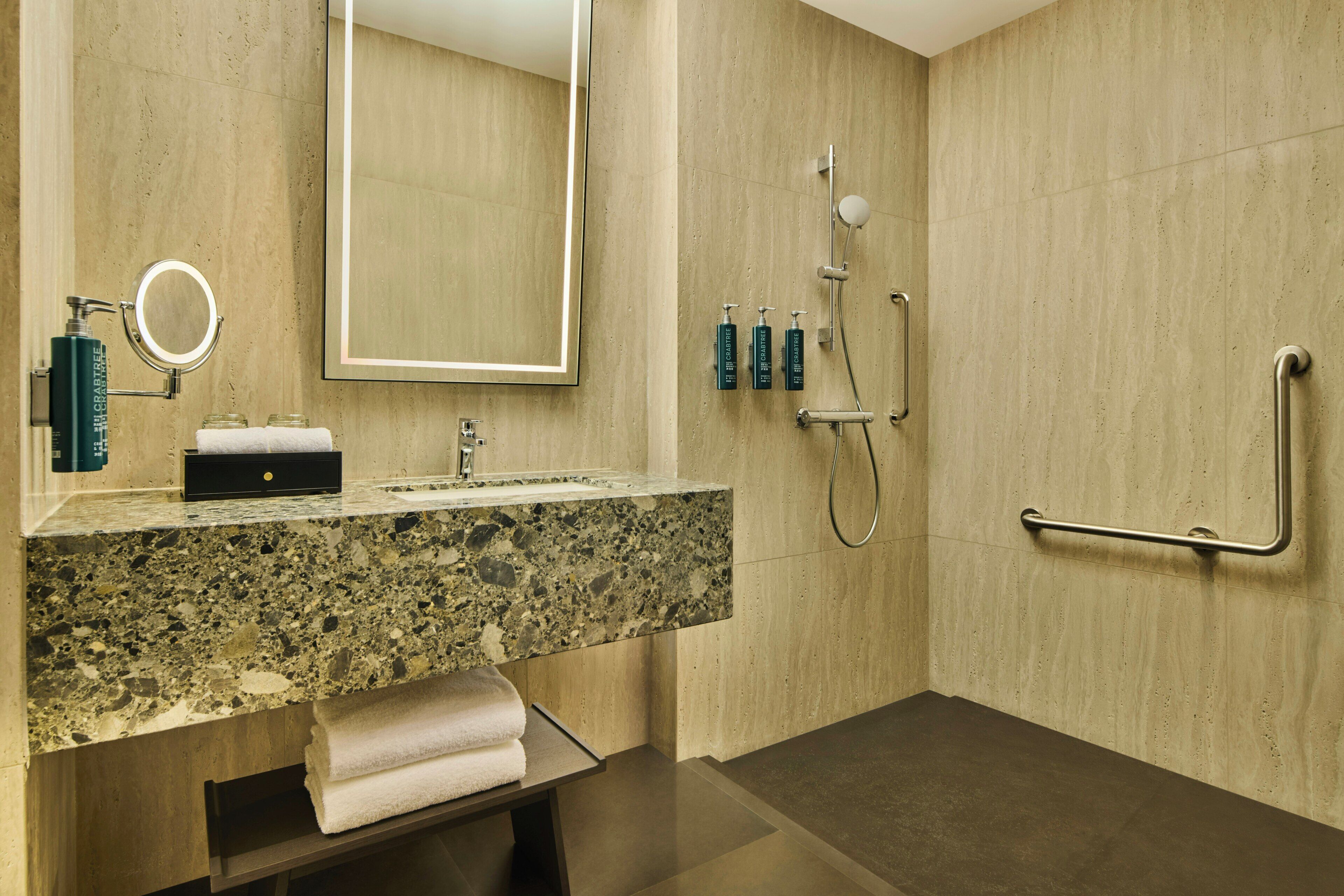 Un baño moderno con encimera de granito, un lavamanos, un espejo, un perchero y una ducha con cabezal de ducha manual, además de una barra de apoyo.
