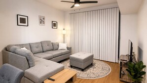 Ein modernes Wohnzimmer mit einer grauen Eckcouch, einem kleinen Hocker, einem Holztisch, einem Fernseher auf einem Schrank und einem Deckenventilator.