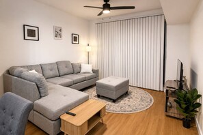 Ein modernes Wohnzimmer mit einer grauen Eckcouch, einem kleinen Hocker, einem Holztisch, einem Fernseher auf einem Schrank und einem Deckenventilator.