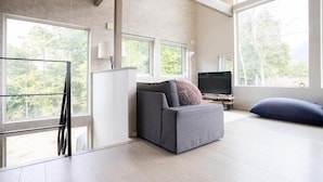 A modern living room with a grey sofa, a television, and large windows offering a view of trees. - GREY HOUSE NISEKO (Niseko)