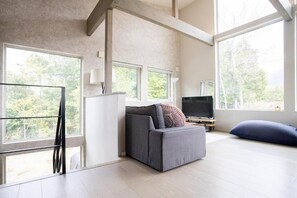 Ein modernes Wohnzimmer mit einem grauen Sofa, einem Fernseher und großen Fenstern mit Blick auf Bäume.