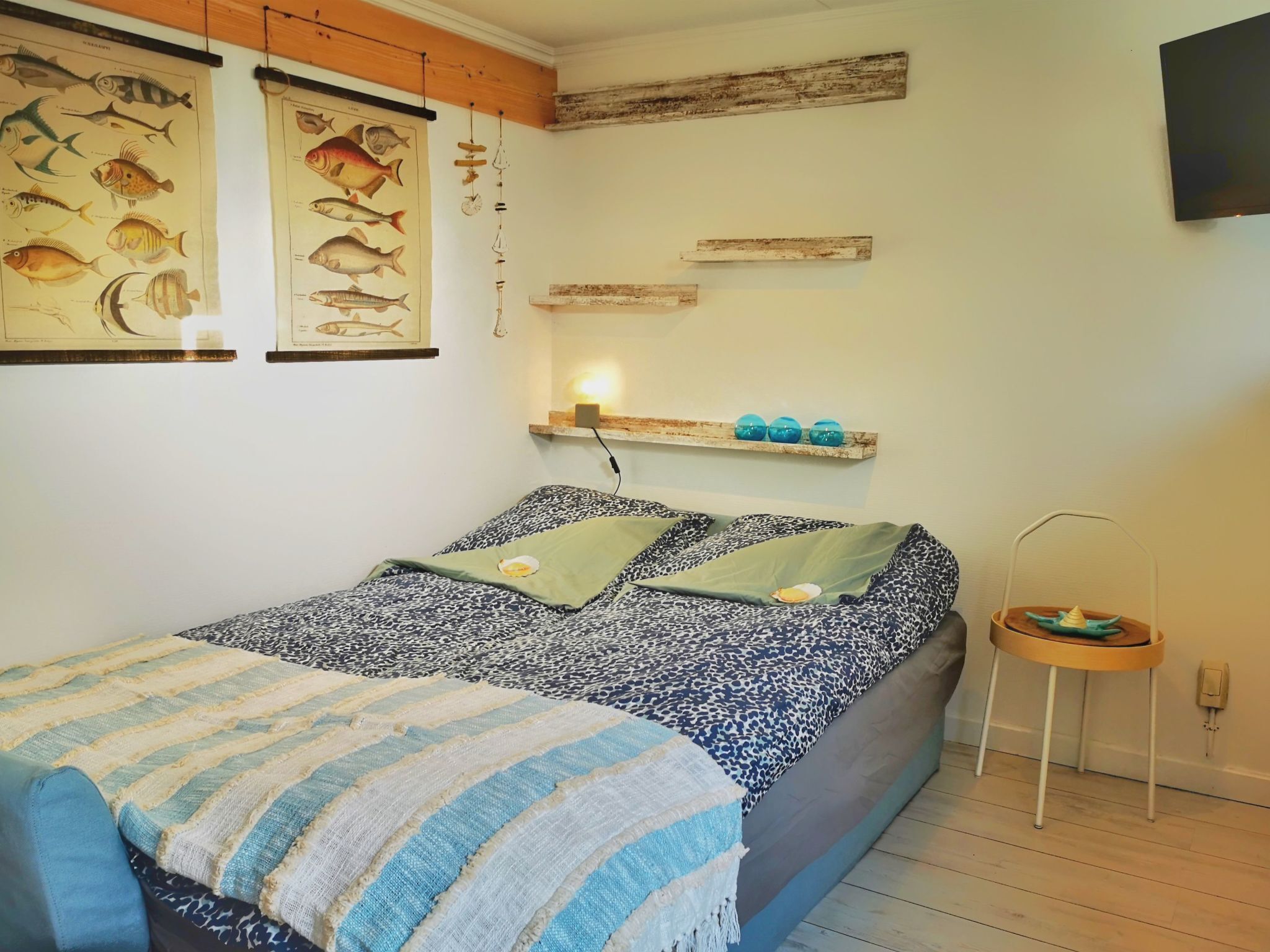 Une chambre à coucher comprenant un lit, une petite table de chevet, une étagère murale décorée d’objets décoratifs et deux œuvres d’art murales sur le thème des poissons.