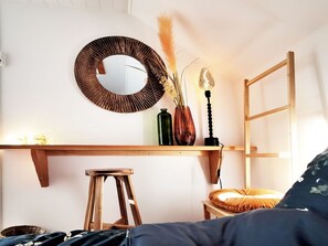 Une chambre à coucher comprenant un lit, un tabouret en bois, un miroir, une étagère avec des objets décoratifs et une table de chevet avec une lampe.