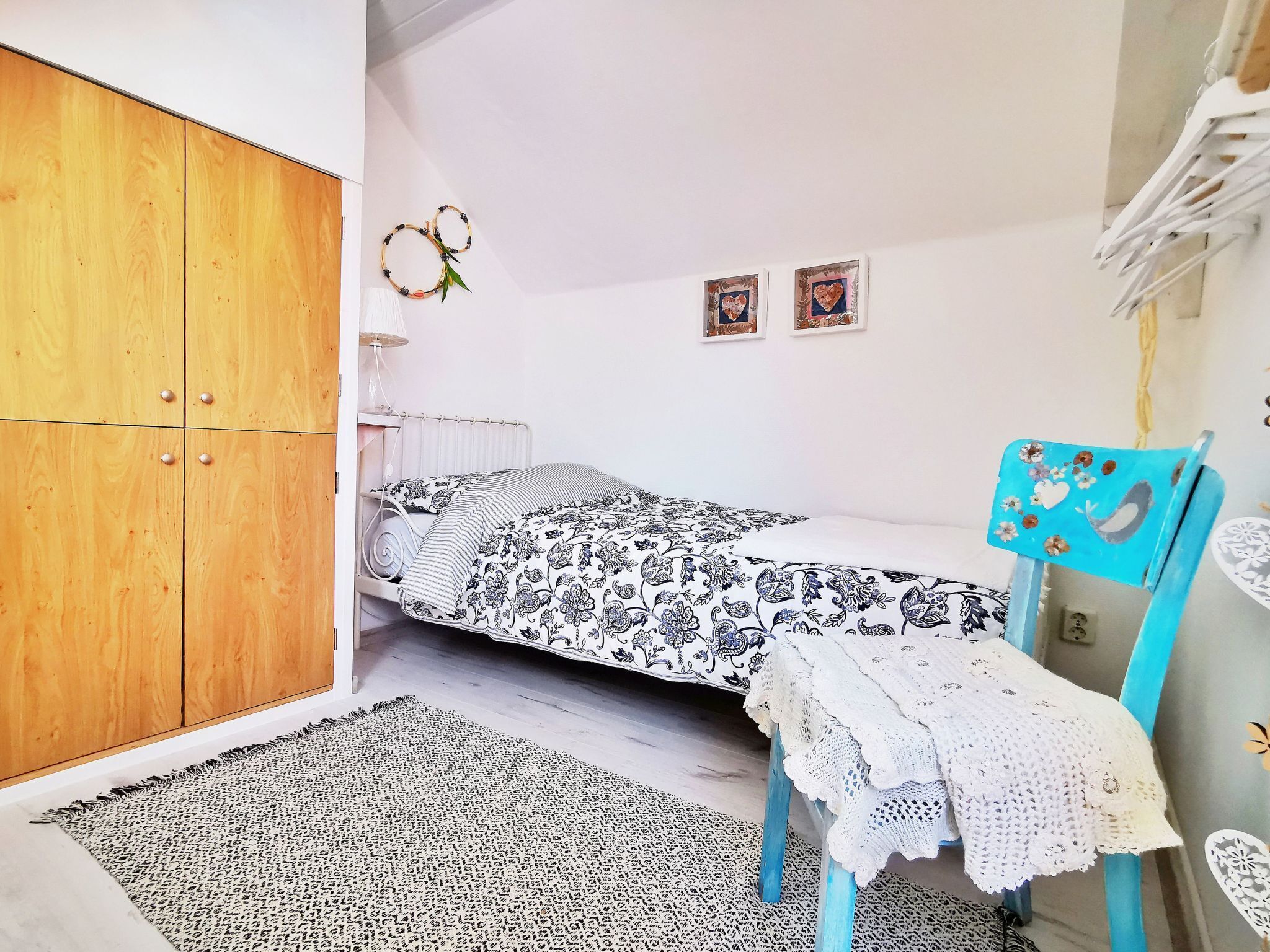 A bedroom with a bed, a wooden wardrobe, a blue chair, a small table, and a wall with two framed pictures.