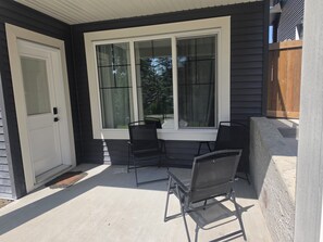 Eine überdachte Terrasse mit Tisch und zwei Stühlen, einer Tür und einem Fenster mit Blick auf Bäume.