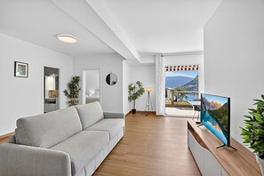 A modern living room with a grey sofa, wooden flooring, a flat-screen TV, and a view of a mountain landscape through a large window. - Lugano Sun & View Apartment for 4, Lugano, Switzerland (Lugano)