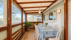 Een overdekt terras met een tafel voor vier personen, witte plastic stoelen, een bloempot en een uitzicht op een huis op de achtergrond.