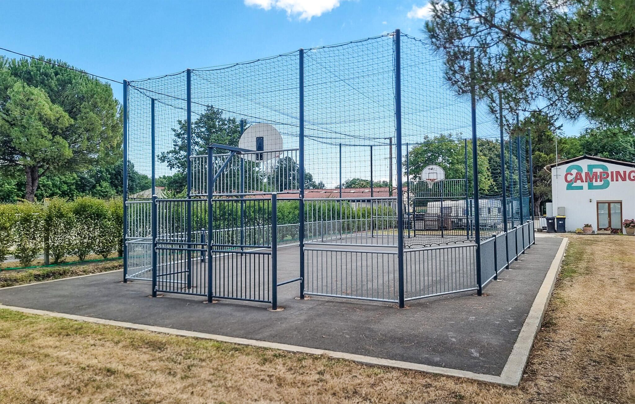 Un terrain de basket-ball entouré de clôtures, avec un panier, bordé d’arbres et à proximité d’un bâtiment portant l’inscription « CAMPING ».