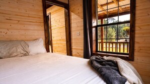 A wooden cabin bedroom with a bed, a window with a view of trees, and a door leading to another room. - The Whole SquatchVilla: Private 4-Cabin Bigfoot Retreat Near Allegheny NF (Marienville)