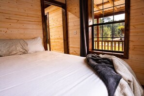A wooden cabin bedroom with a bed, a window with a view of trees, and a door leading to another room. - The Whole SquatchVilla: Private 4-Cabin Bigfoot Retreat Near Allegheny NF (Marienville)