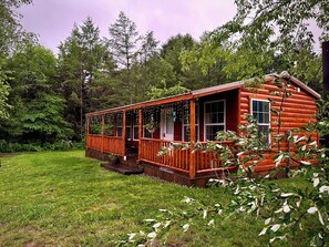 A log cabin with a porch, surrounded by a grassy yard and dense forest. - The Whole SquatchVilla: Private 4-Cabin Bigfoot Retreat Near Allegheny NF (Marienville)