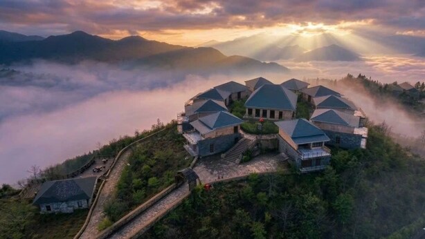 A cluster of traditional-style buildings with steep roofs, surrounded by lush greenery and a mountainous backdrop.