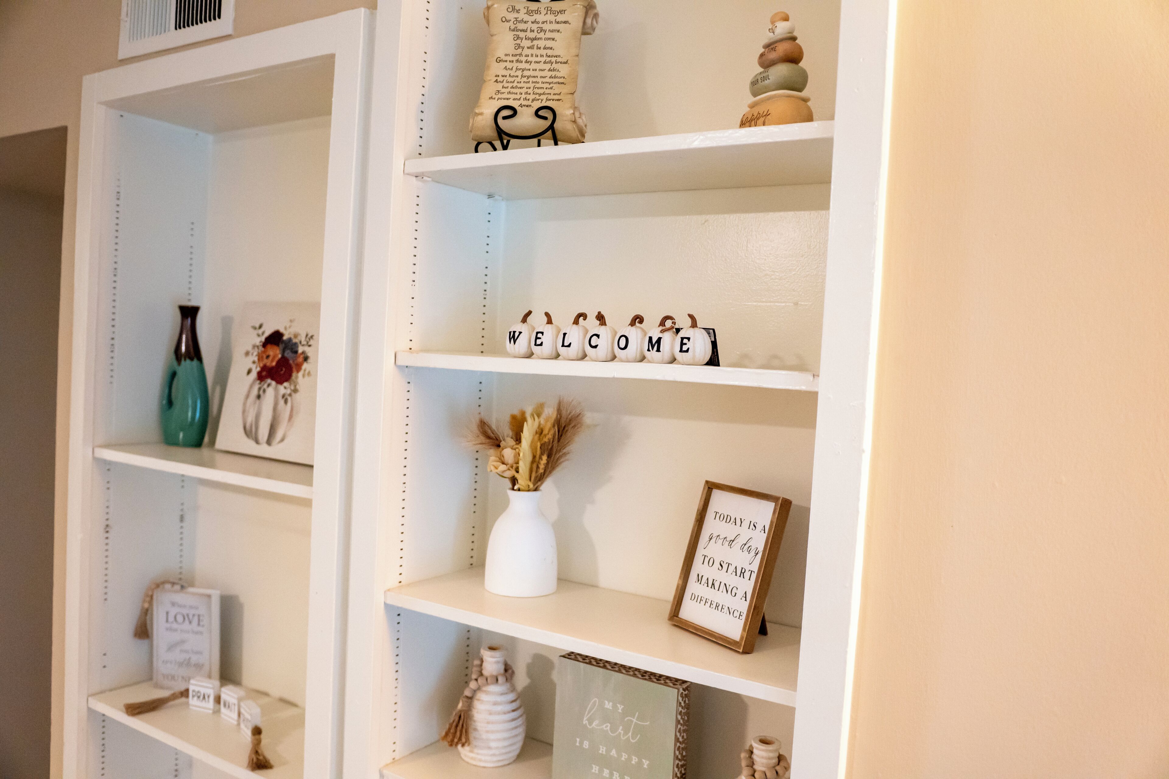 A white shelf with decorative items including a 'WELCOME' sign, a framed quote, and small vases.