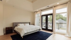 A bedroom with a bed, a dark rug, wooden flooring, a nightstand, and large windows with curtains.