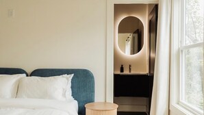 A bedroom with a bed, a round side table, a mirror, and a view of a bathroom.