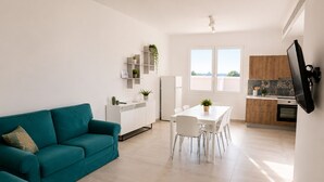 A modern living space with a teal sofa, a dining table with chairs, a kitchen area with wooden cabinets, and a flat-screen TV mounted on the wall. - Camera Osanna by Symphonya Rentals (Nardò)