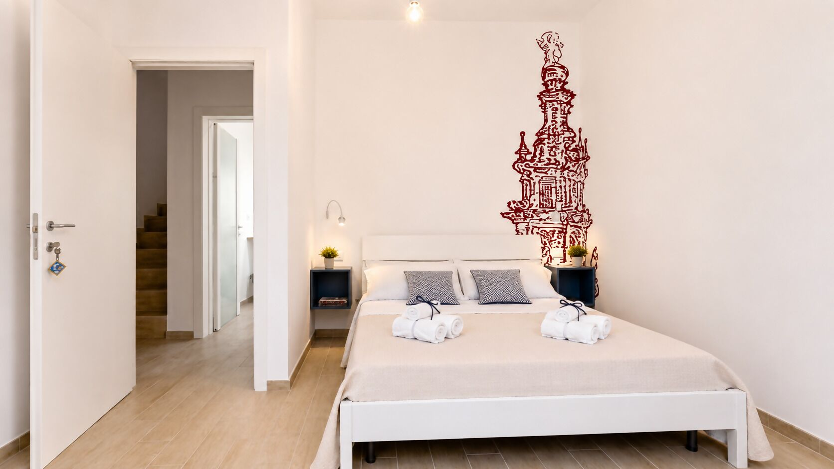 A modern hotel room with a bed, bedside tables, a nightstand, a decorative wall piece, and a staircase in the background.