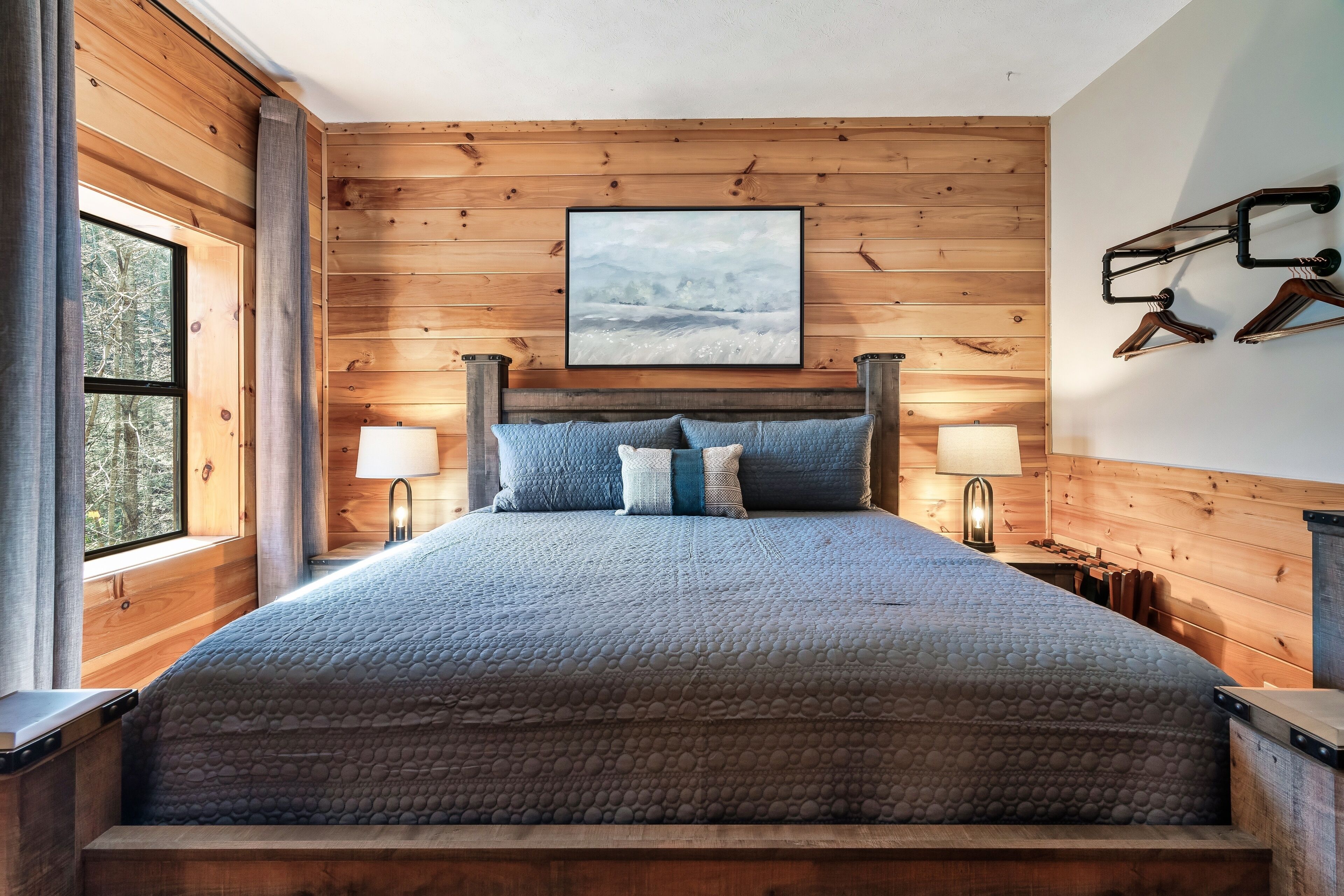 Ein Schlafzimmer mit Holzwänden, einem Bett, zwei Nachttischlampen, einem gerahmten Bild und einem Fenster mit Vorhängen.