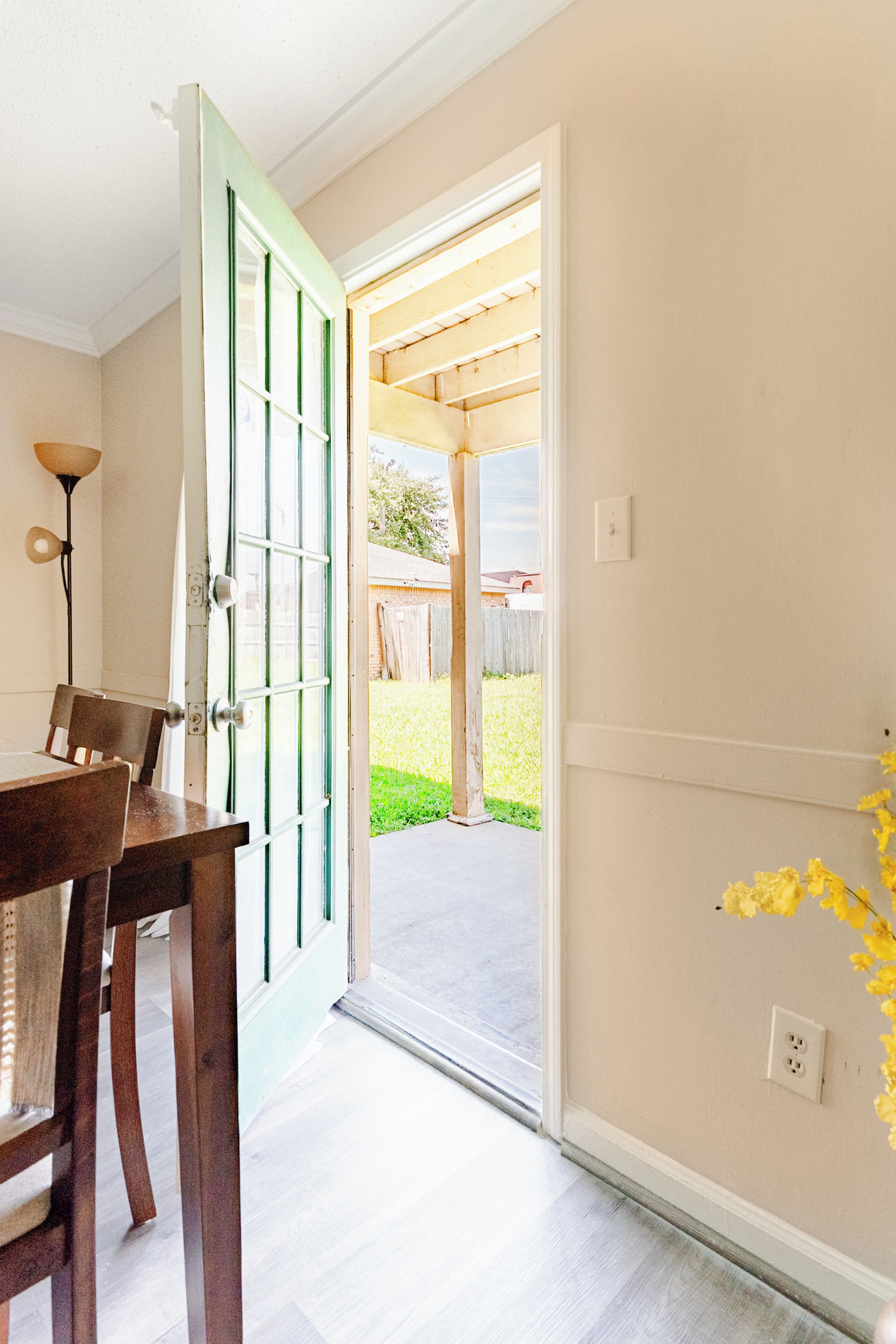 An interior space with a wooden table and chairs, a green-framed door leading outside, and a white wall with a power outlet.