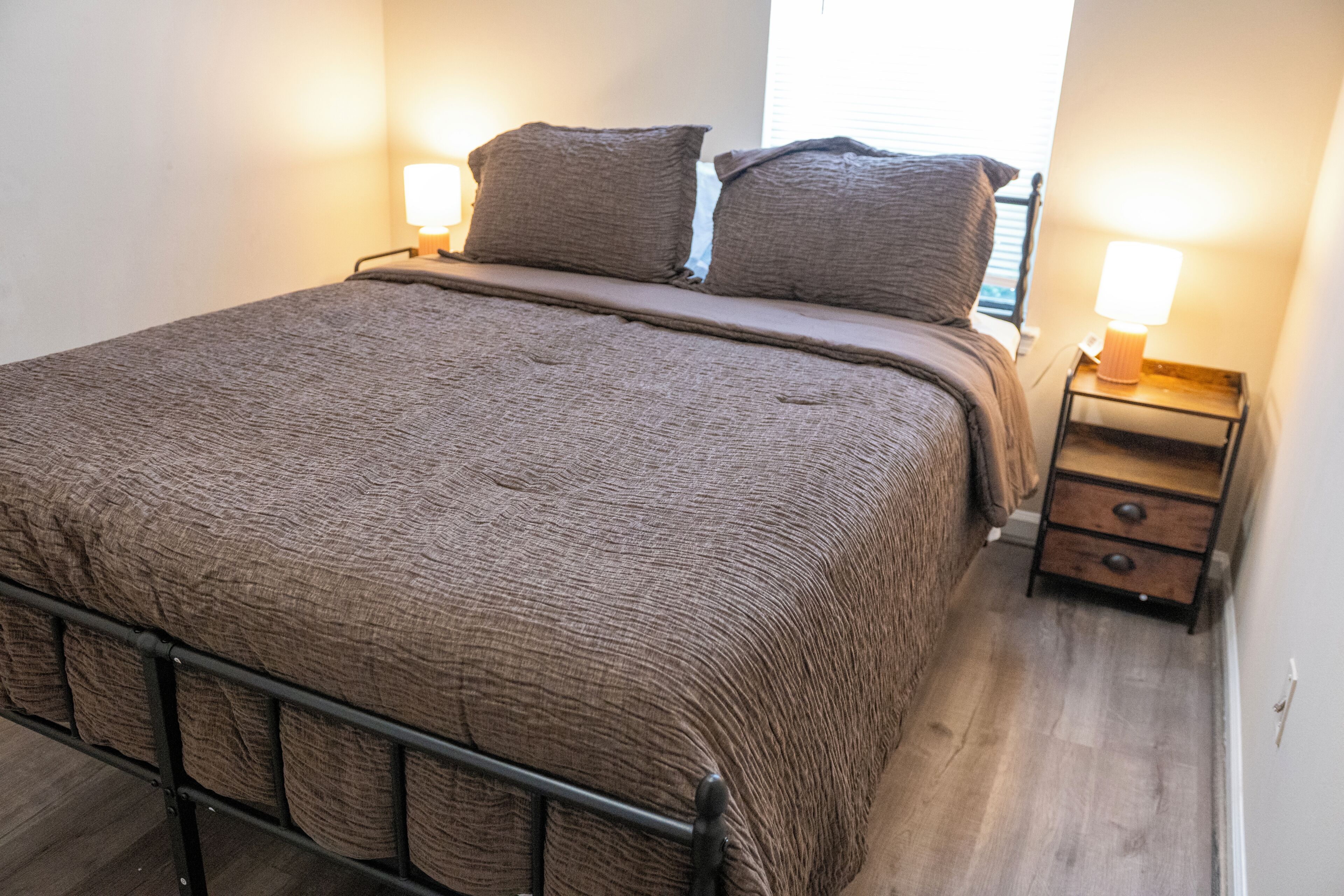 Ein ordentlich bezogenes Bett mit Metallgestell, zwei Nachttische mit Lampen und ein Holzboden.