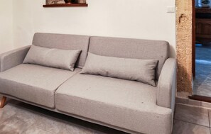 A grey sofa with two cushions in a room with a tiled floor and a wooden shelf above. - Gorgeous home in Sernhac (Sernhac)