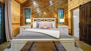 A bedroom with a sloped ceiling, wooden walls, a bed with a headboard, bedside tables, and curtains. - Gnarly Ever After | Luxury Meets Fun (Broken Bow)
