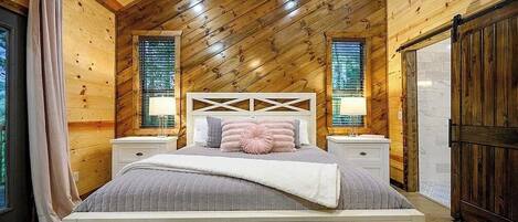 Un dormitorio con techo inclinado, paredes de madera, una cama con cabecera, mesitas de noche y cortinas.