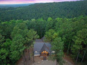 A cabin surrounded by dense forest, with a sloped roof and a central entrance. - Gnarly Ever After | Luxury Meets Fun (Broken Bow)