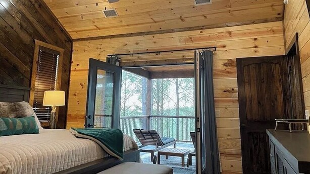 A wooden cabin bedroom with a bed, a desk, a chair, and a balcony with a view of trees. - Gnarly Ever After | Luxury Meets Fun (Broken Bow)